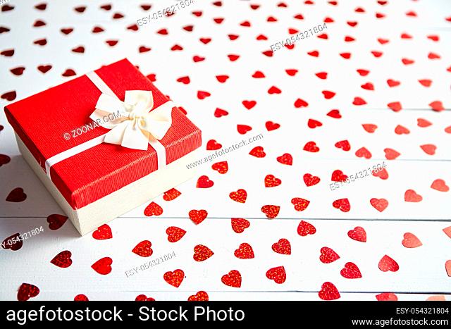 Valentine's Day decoration composition. Boxed gift placed on heart shaped red sequins on white wooden table. Romantic background. Flat lay, top view