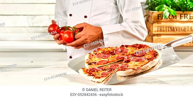 Chef with a fresh oven-fired Italian pepperoni pizza on a metal paddle standing in the background holding fresh tomatoes in his hands