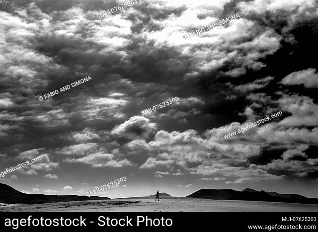 Dramatic dark sky with clouds above a single man walking in the desert. Mountains far in a beautiful landscape