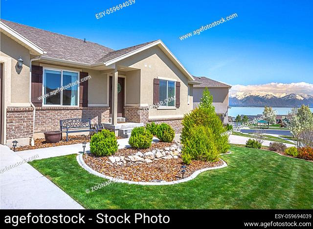 Home with landscaped garden overlooking Utah lake and snowy Mount Timpanogos. Brown front door, windows with shutters, and brick wall can also be seen at the...