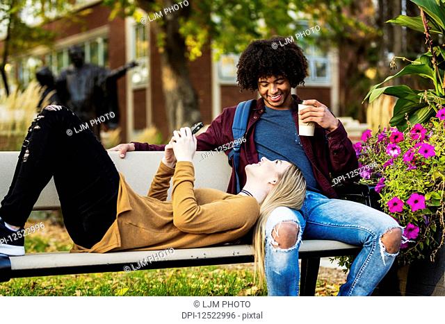Young dating couple who are university students relaxing on a bench on campus and the woman is checking her smart phone while laying her head on her boyfriend's...