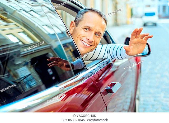 Nice to see you. Cheerful positive adult man smiling and waving to his friend while putting his hand out of the window