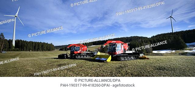 16 March 2019, Baden-Wuerttemberg, Schonach: Two snow groomers are standing in front of a windmill on a green meadow. For many lift operators in...