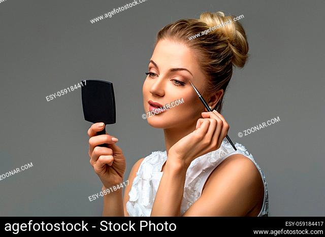 beautiful blond young woman with hairdo holding pocket mirror and applying makeup. studio beauty shot. copy space