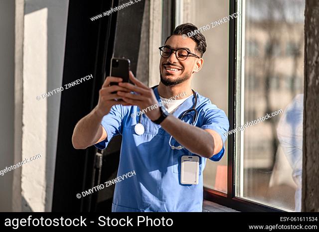 Waist-up portrait of a smiling pleased handsome physician in eyeglasses photographing himself with the smartphone