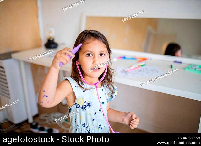 Picture a little brunette girl playing with a purple toy thermometer and a pink toy stethoscope, with a blurry mirror and a white desk with coloring images and...