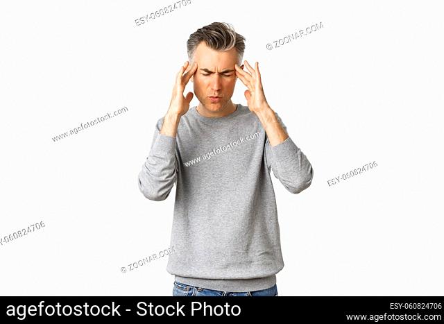 Portrait of middle-aged man in grey sweater having a headache, touching head and grimacing from painful migraine, standing over white background