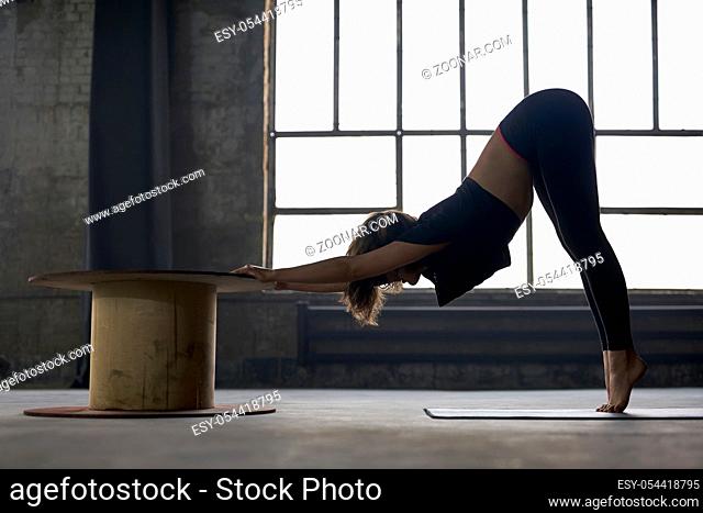 Cute girl in a black sportswear engaged in yoga on the window background in a loft style hall. She stands on the toes on the mat and does a forward tilt while...