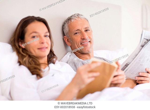 Happy woman reading a book while her husband is reading the news in their bedroom
