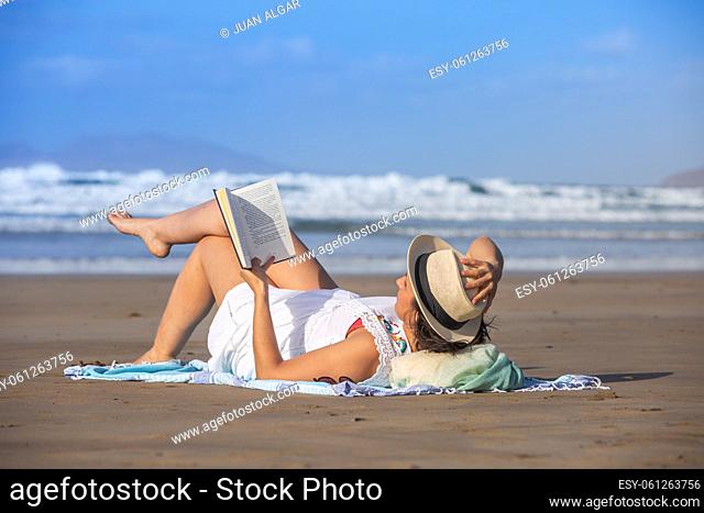 Female traveler touching hat and enjoying reading while lying on sand against waving sea and blue sky on summer weekend day on resort