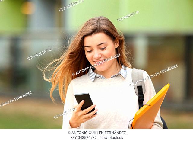 Single student walking and reading mobile phone messages with a university building in the background