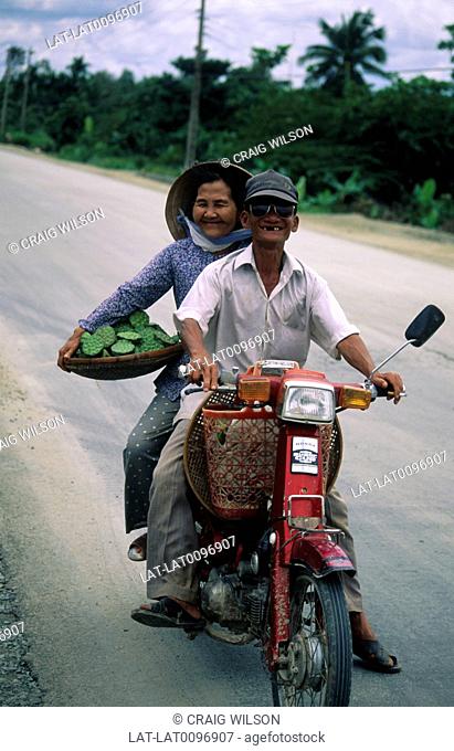 A man driving a motor scooter, and a woman pillion passenger holding a tray of lotus leaves