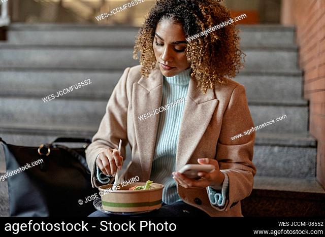 Young woman holding mobile phone having food on staircase