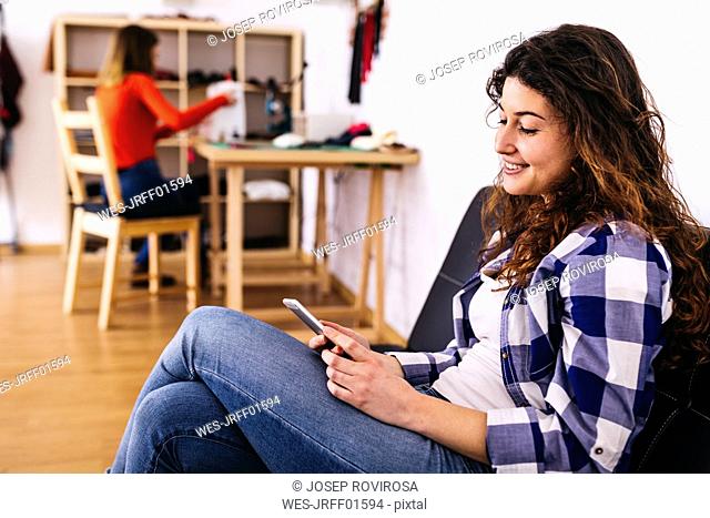 Smiling young woman in fashion studio using cell phone with woman in background on sewing machine