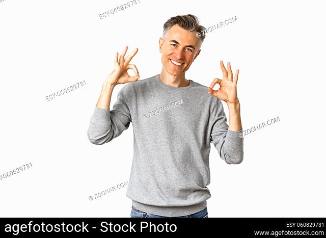 Portrait of successful handsome man in grey sweater, showing okay signs and smiling satisfied, approve something good, recommending product