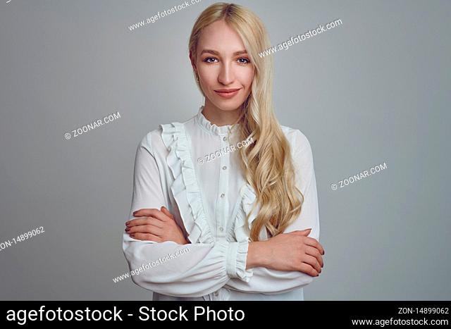 Young and beautiful long-haired blond woman in white blouse standing with her arms folded and looking at camera with a smirk