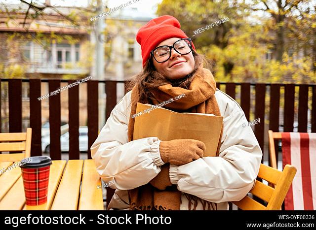 Woman in warm clothing embracing book while sitting at cafe