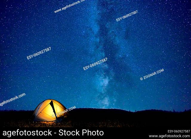 Illuminated tent in mountains under night dark blue sky with many stars