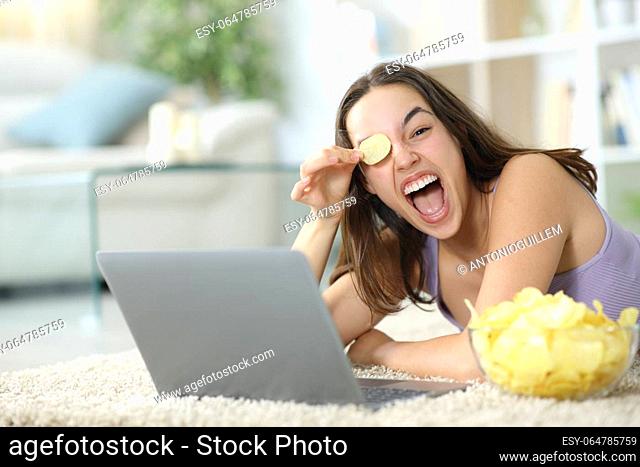 Happy woman with laptop playing with potato chips lying on the floor at home