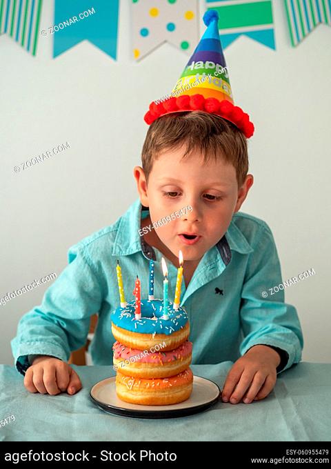 Five year old boy blows out the candles on festive donuts cake in his birthday. Happy little child and plate with doughnuts stack cake with candles on the table