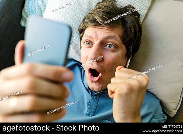 Excited man holding smart phone lying down on bed at home