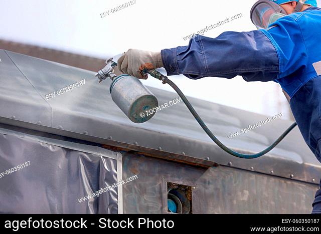Industrial work. Priming of metal products from the compressor gun. A worker in overalls and a protective mask paints the body of a truck trailer or a metal car