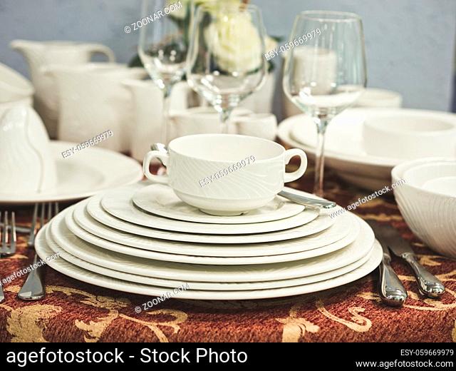 Set of new dishes on table with tablecloth. Stack of white plates and wine glasses with flowers on restaurant table. Shallow DOF
