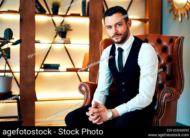Handsome young man sitting and posing in armchair in modern luxury interior. Fashion portrait