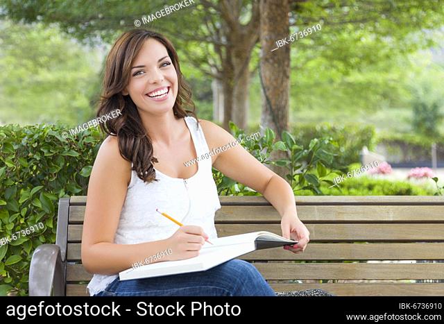 Attractive young adult female student on bench outdoors with books and pencil