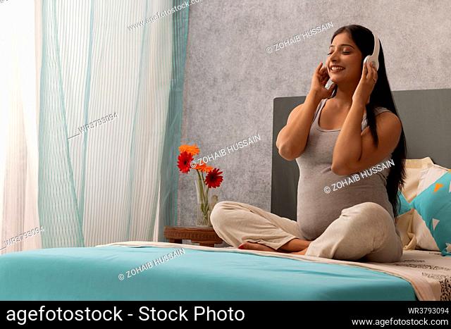 Pregnant woman listening to music with headphone while sitting on bed at home