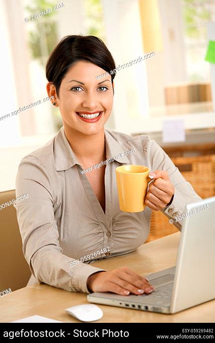 Happy young woman drinking tea at desk, using laptop computer, smiling