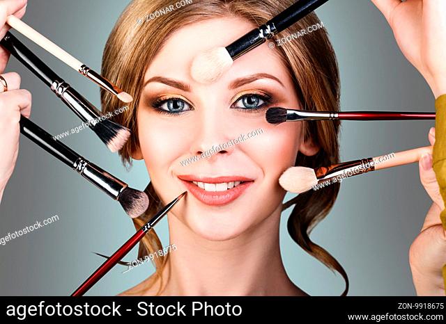 Many hands with cosmetics brushes doing make-up to glamour woman, over gray background