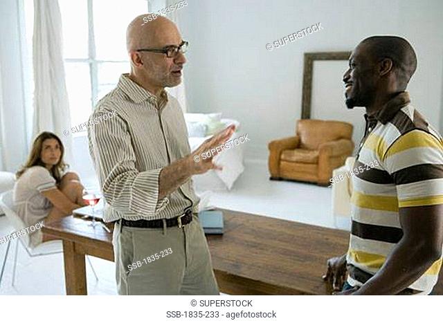 Mature man talking to a mid adult man with a woman sitting on a chair in the background