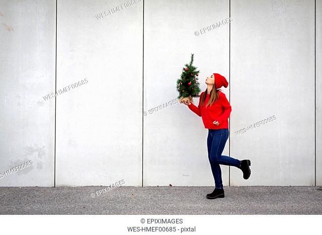 Woman wearing red pullover and wolly hat, holding artificial Christmas tree in front of a wall