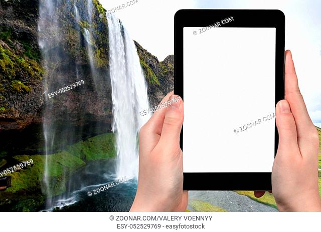 travel concept - tourist photographs Seljalandsfoss waterfall of Seljalands River in Katla Geopark in Iceland in autumn on tablet with cut out screen for...