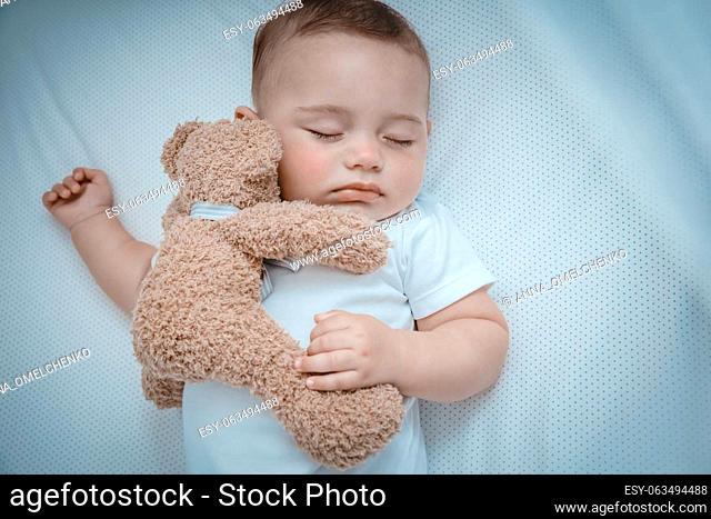 Closeup photo of a cute little baby boy napping at home in the crib with a soft toy bear. Sweet child sleeping. Happy healthy family life