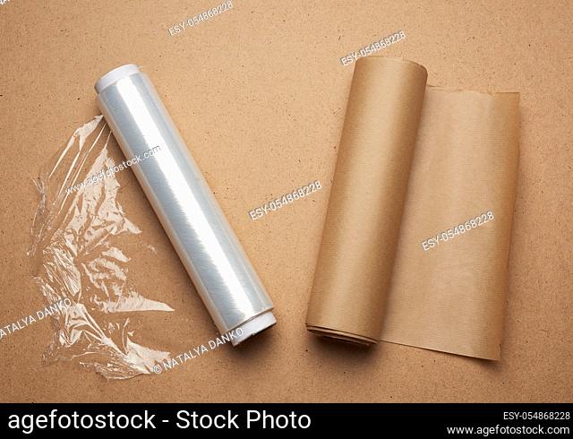 big roll of wound white transparent film for wrapping food and roll of brown craft paper, brown wooden background. rejection of plastic packaging in favor of...
