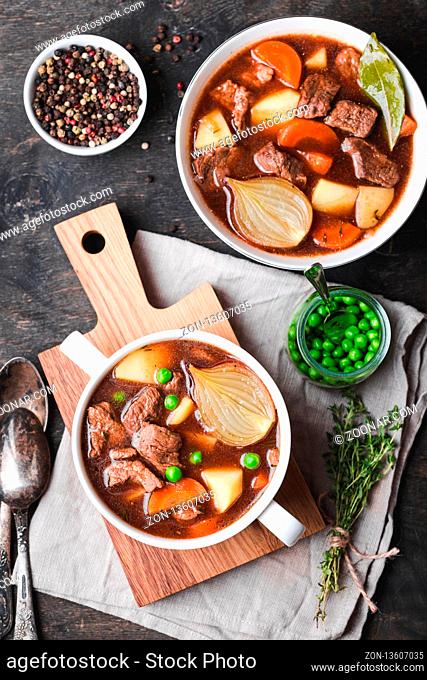 Meat stew with beef, potato, carrot, pepper, spices, green peas. Slow cooked meat stew, bowl, wooden background. Hot autumn/winter dish. Closeup