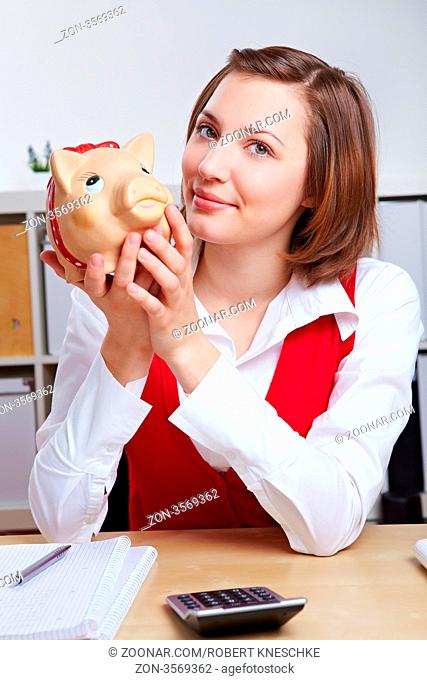 Attraktive Frau sitzt mit Sparschwein am Schreibtisch im Büro