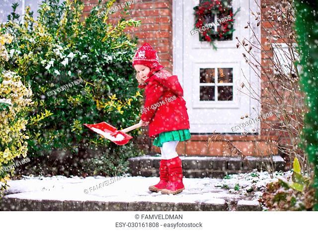 Little girl shoveling snow on home drive way. Beautiful house decorated for Christmas. Child with shovel playing outdoors in Xmas season