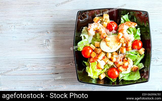 top view of Cesar salad with prawns in black bowl on gray wooden table with panoramic copyspace