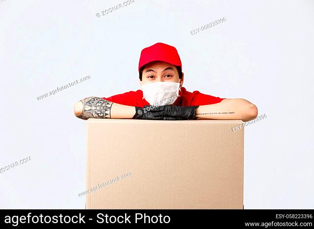 Delivery, online shopping and quarantine concept. Cheerful asian delivery guy in red cap uniform, medical mask and protective gloves, leaning on large box