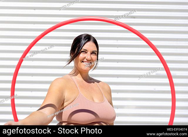 Happy curvy woman looking through plastic hoop standing in front of corrugated wall