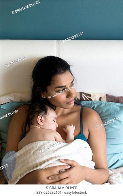 Young mother lying in bed with her baby son asleep on her chest. They both have a blanket over them and the mother is looking to the side