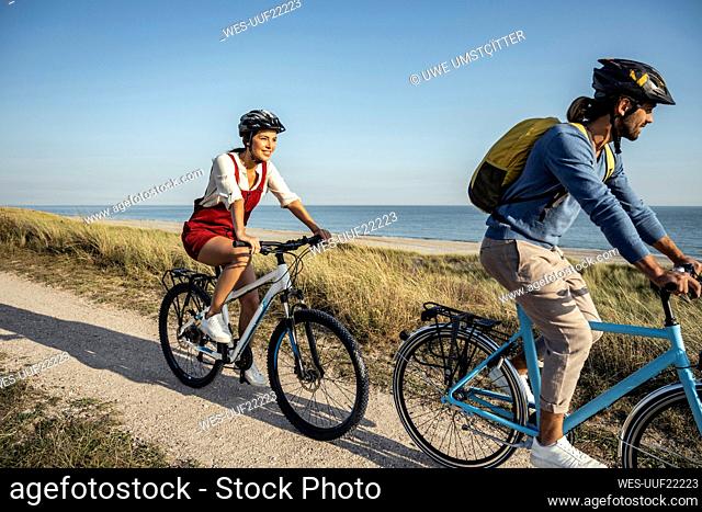 Young woman smiling while riding bicycle with man on footpath against sea