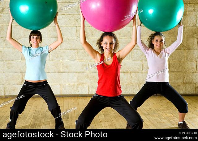 A group of women making exercise holding big balls over heads and standing astride. They're smiling and looking at camera. Front view