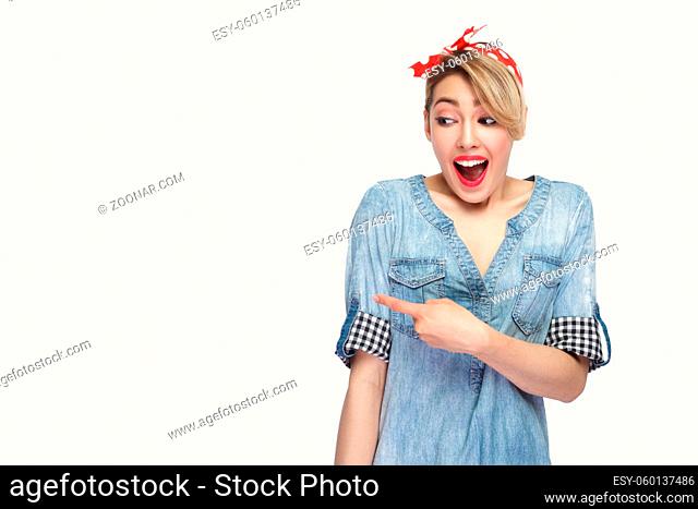 Portrait excited of beautiful young woman in casual blue denim shirt with makeup, red headband standing looking and pointing at background copyspace