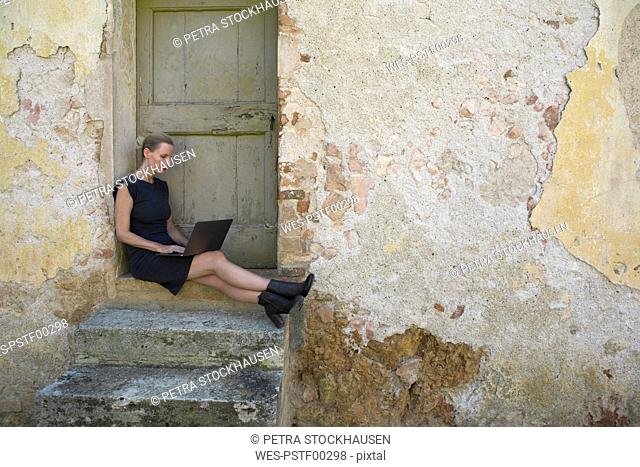 Italy, Tuscany, Monteriggioni, woman sitting at house entrance using laptop