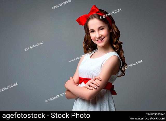 Beautiful teenage girl with long curly hair and red ribbon bow on head wearing white dress. Happy expression. Studio portrait on grey background