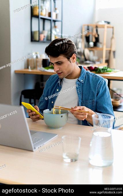 Young man using smart phone while having noodles at home